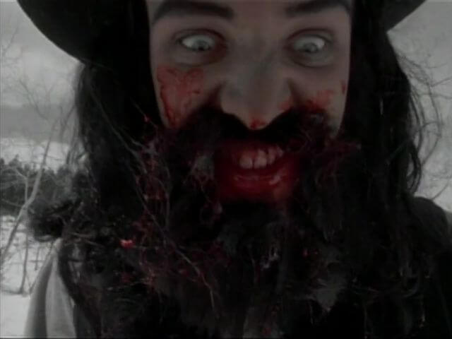 A close-up shot of a wild-eyed man with a prospector's hat, a thick black beard, and bloody mouth.