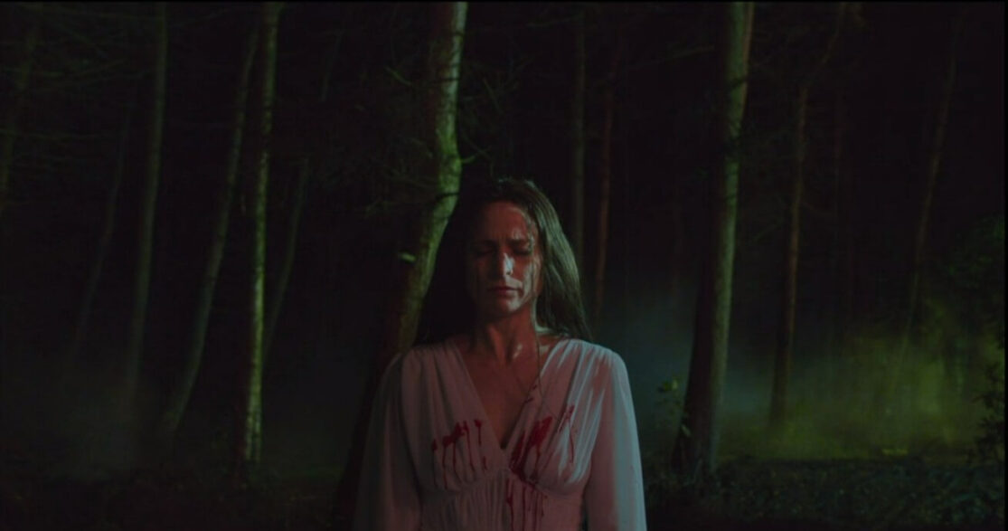 A bloody woman in a gown stands in a forest.