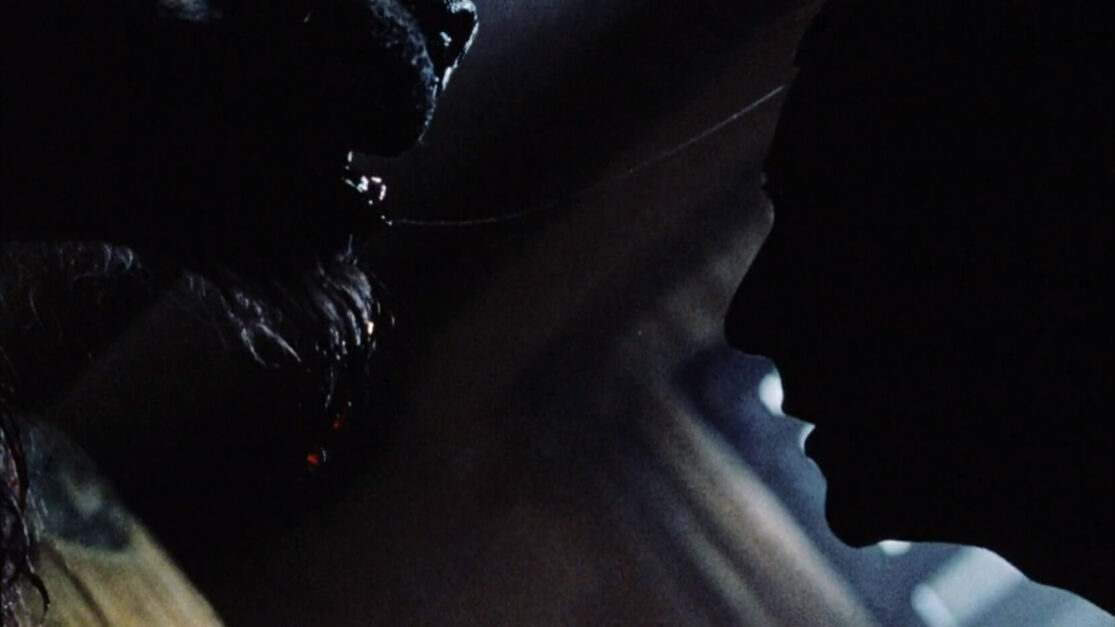 In a dark room, the silhouetted profile of a man faces that of a drooling werewolf.