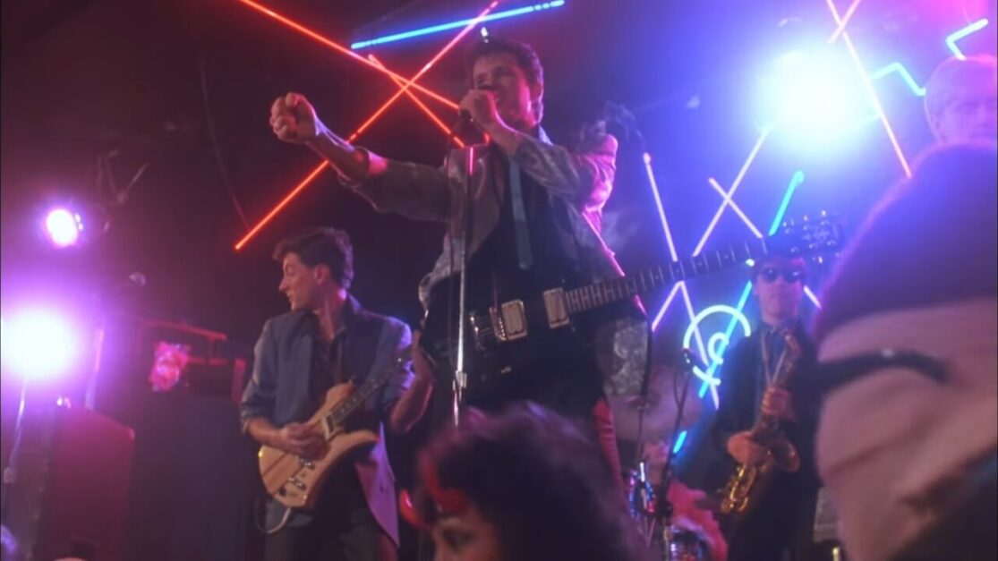 A new wave band performs in front of a neon backdrop.