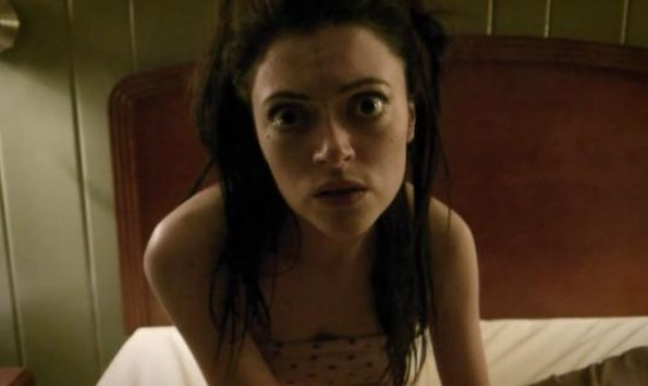 A young woman in a strapless dress sits on a hotel bed. She appears to be speaking to someone off-camera.