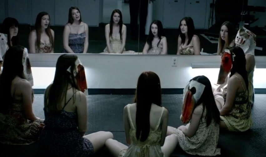 Young girls, possibly cult members, sit in a semi-circle before a mirror, creating the illusion that there's a full circle of girls in the room.