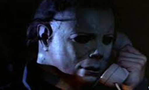 In a dark room, a man in a creepy white mask holds a phone to his ear.