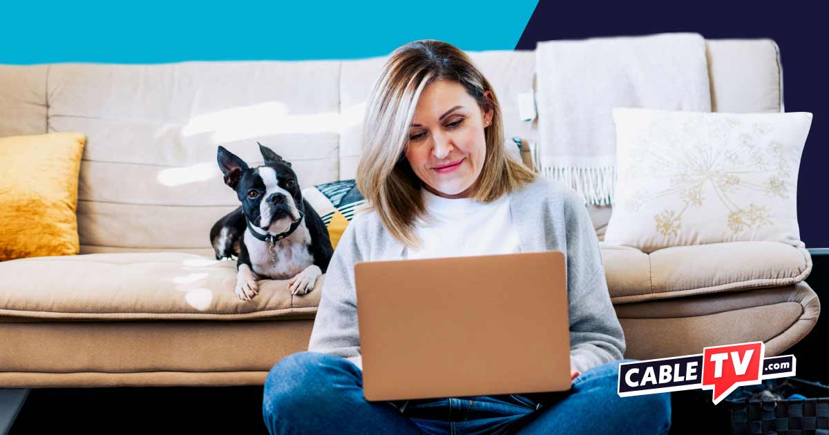 A woman sitting on the floor next to a couch using her computer, and a Frenchie dog next to her