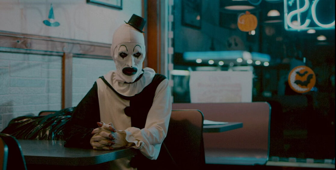 Art the Clown, dressed in black and white, sits at a pizza parlor booth glaring at someone offscreen.