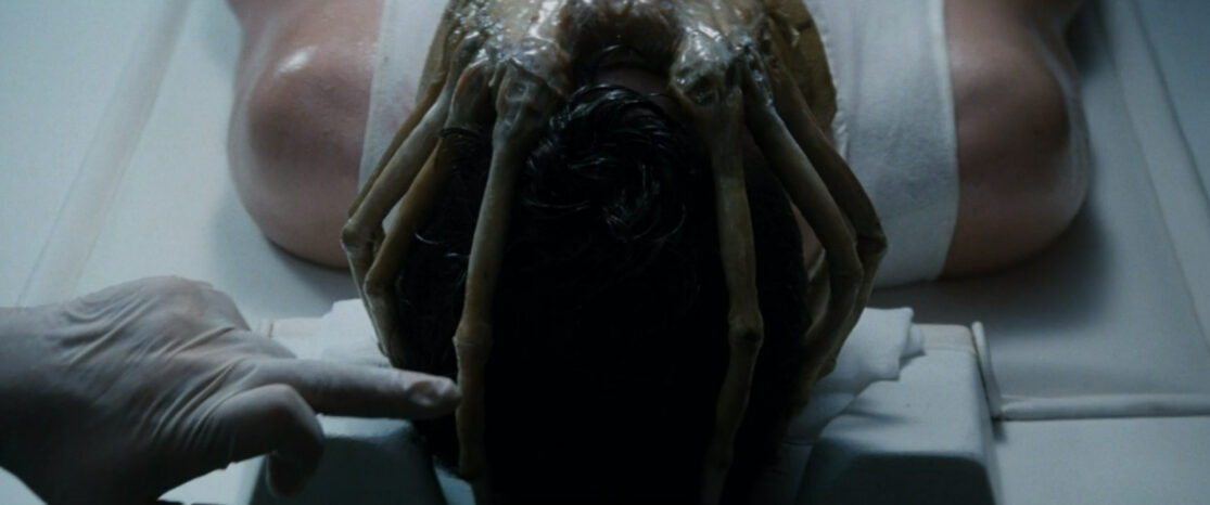 A facehugger alien covers the face of a man lying on an exam table while a gloved hand points at one of the creature's arms.