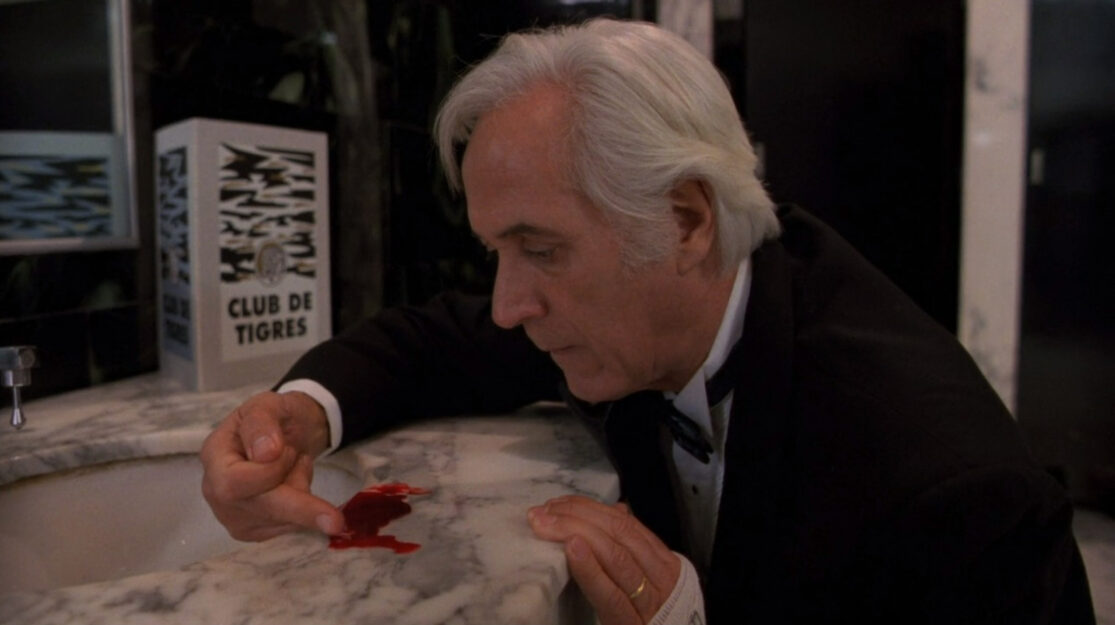 A gray-haired man in a suit uses his finger to gather blood from a bathroom sink counter.
