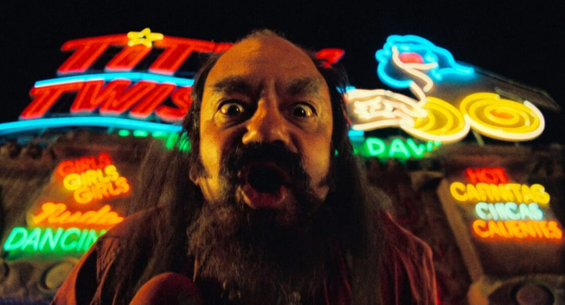 A wild-eyed, hairy man—standing in front of a huge, neon-adorned roadhouse bar, bellows into the camera.