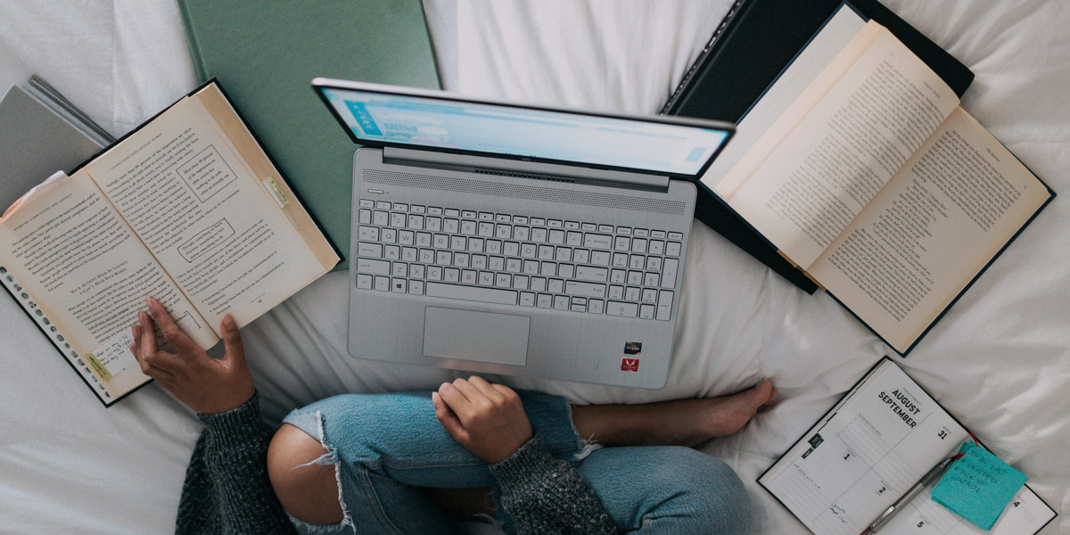 Photo of student crossed legs on bed with laptop and books around them. Photo by Windows on Unsplash