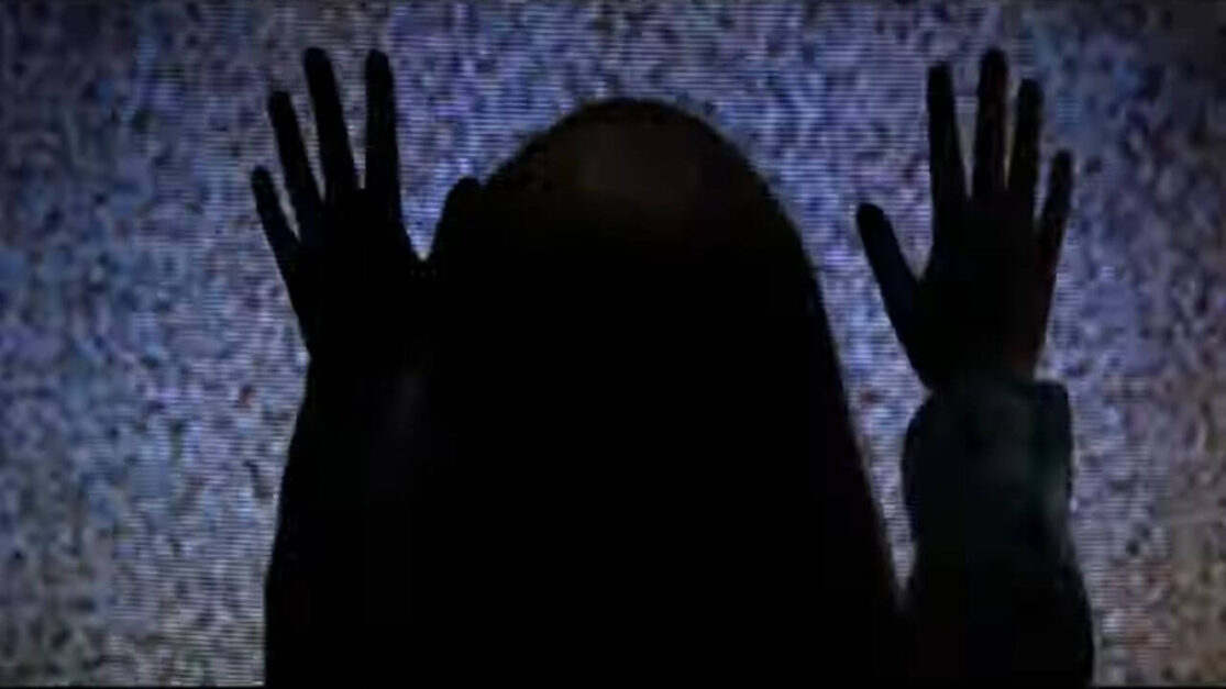 A little girl, her back to the camera, is silhouetted by the TV light. Both of her hands are on the TV screen, which shows only white noise.