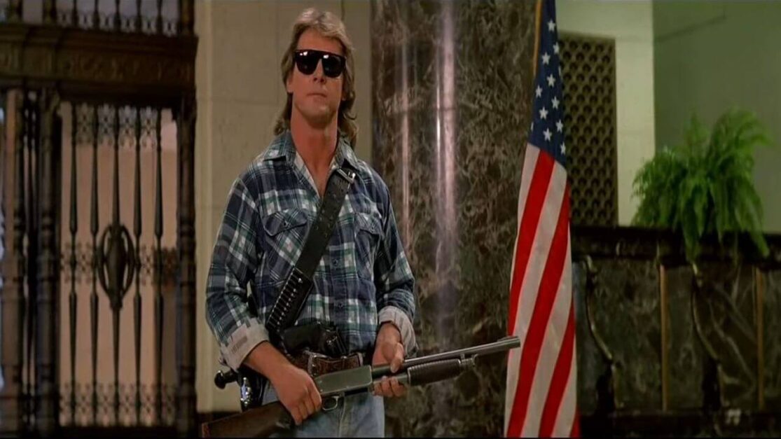 A man in a flannel shirt and sunglasses holds a rifle