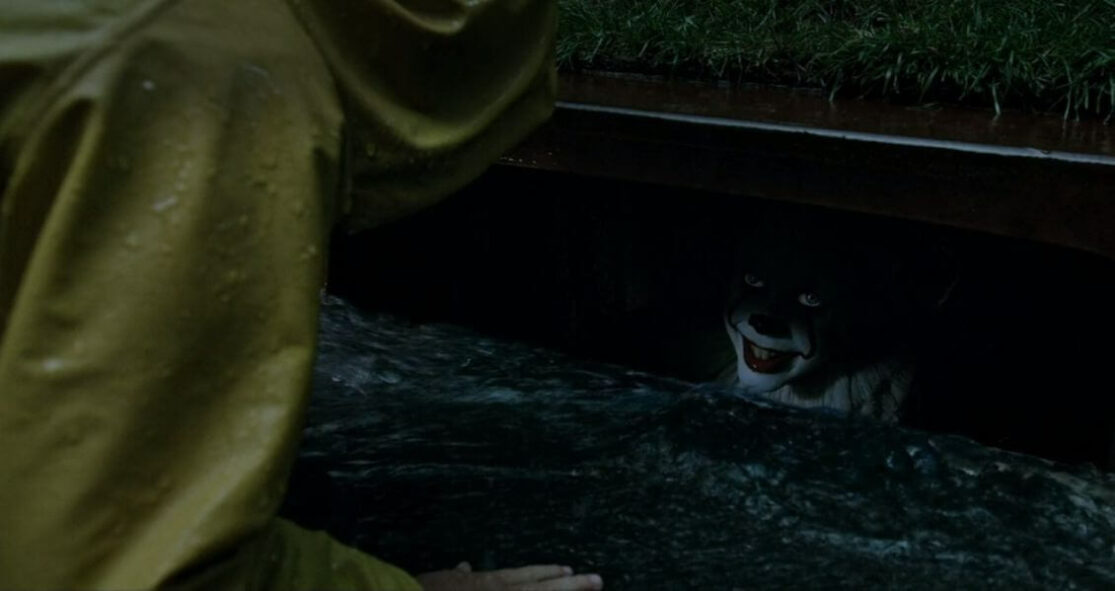 A clown in a storm drain leers up at a young boy in a raincoat.