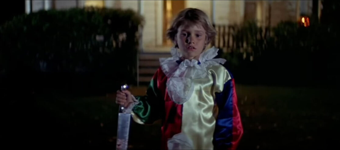 A catatonic young boy dressed as a clown holds a bloody butcher knife outside his home at night.