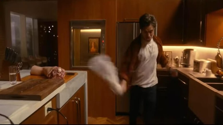 A man dances in his kitchen, twirling a towel, while a human leg rests on a cutting board.