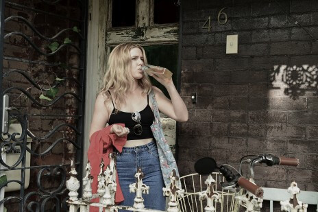 A young woman drinks from a liquor bottle on the porch of an old house.