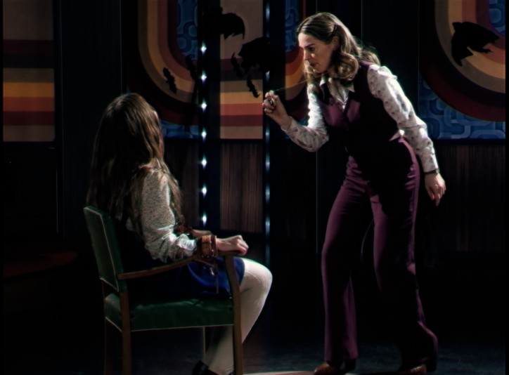 On a soundstage, a woman speaks sternly to a young girl in a chair.