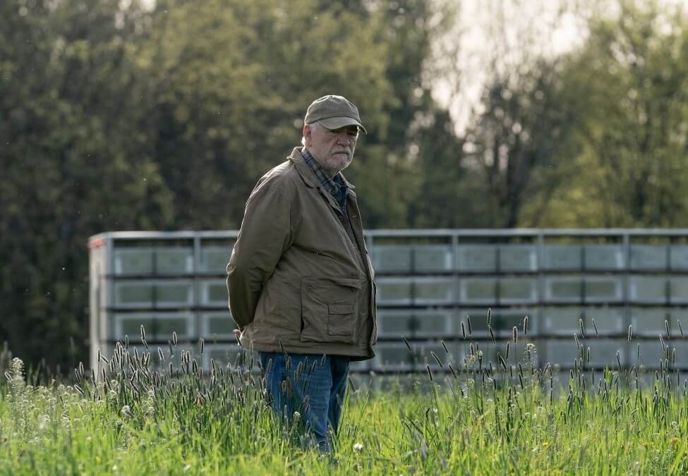 An older man stands in a field looking thoughtful.