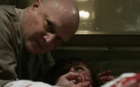 A glaring bald man leans over a bloody woman giving birth.