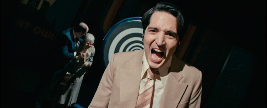 In a scene from late Night with the Devil, a man in a suit appears to scream at the camera while, in the background, a different man consoles a woman as they stand near a hypno-wheel.