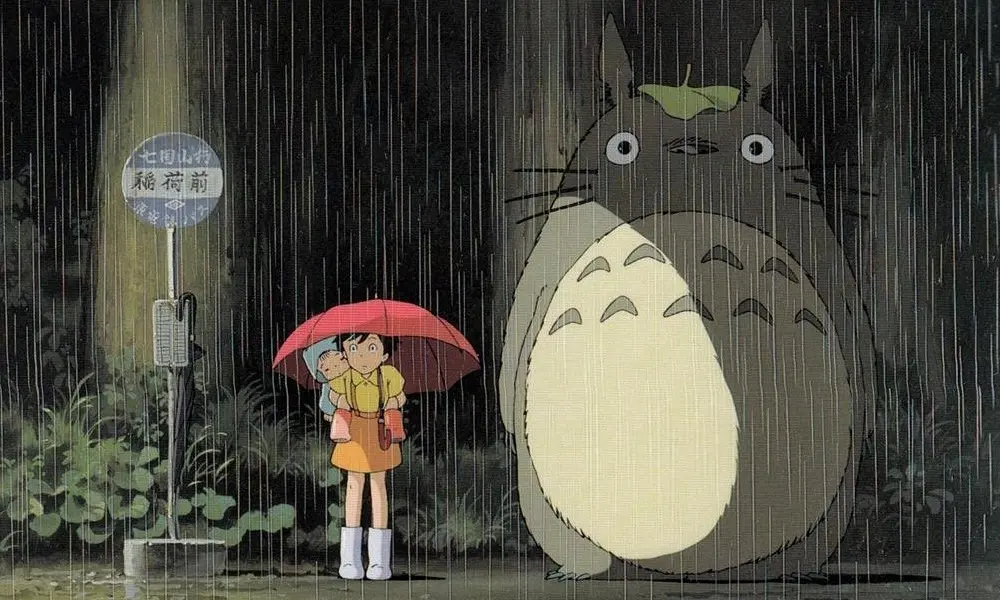 Totoro, a large grey furry creature with a leaf on his head, stands next to a little kid in the rain.