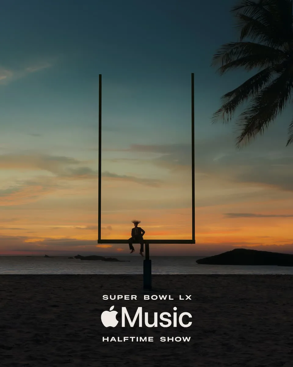 Bad Bunny in a brown suit and traditional pava hat, sitting on a yellow goalpost on a sunny tropical beach.