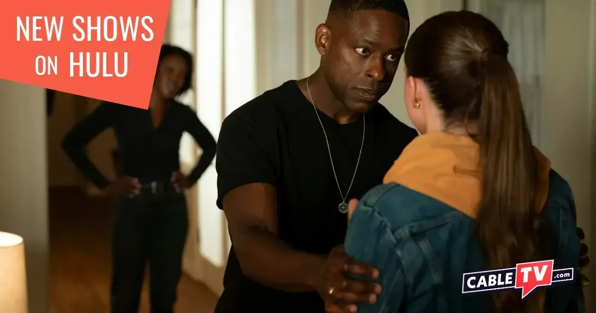 A man holds a young woman by the arms while giving her a serious, paternal look. In the background, the man's wife looks on.