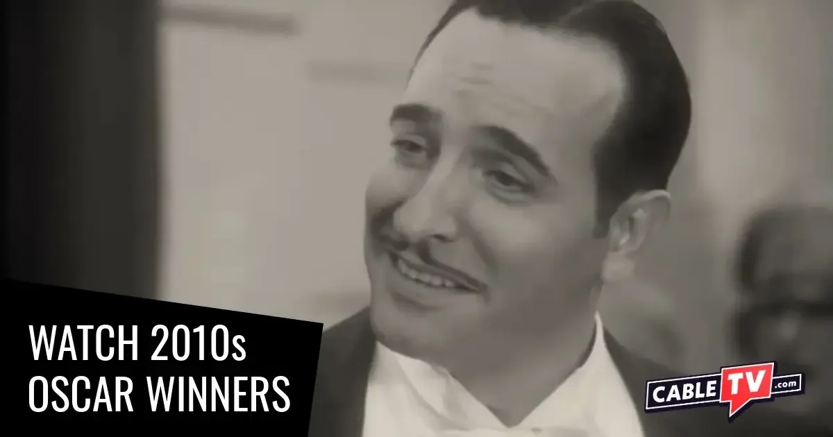 A black-and-white close-up shot of a smiling, dapperly dressed man with a pencil-thin mustache.