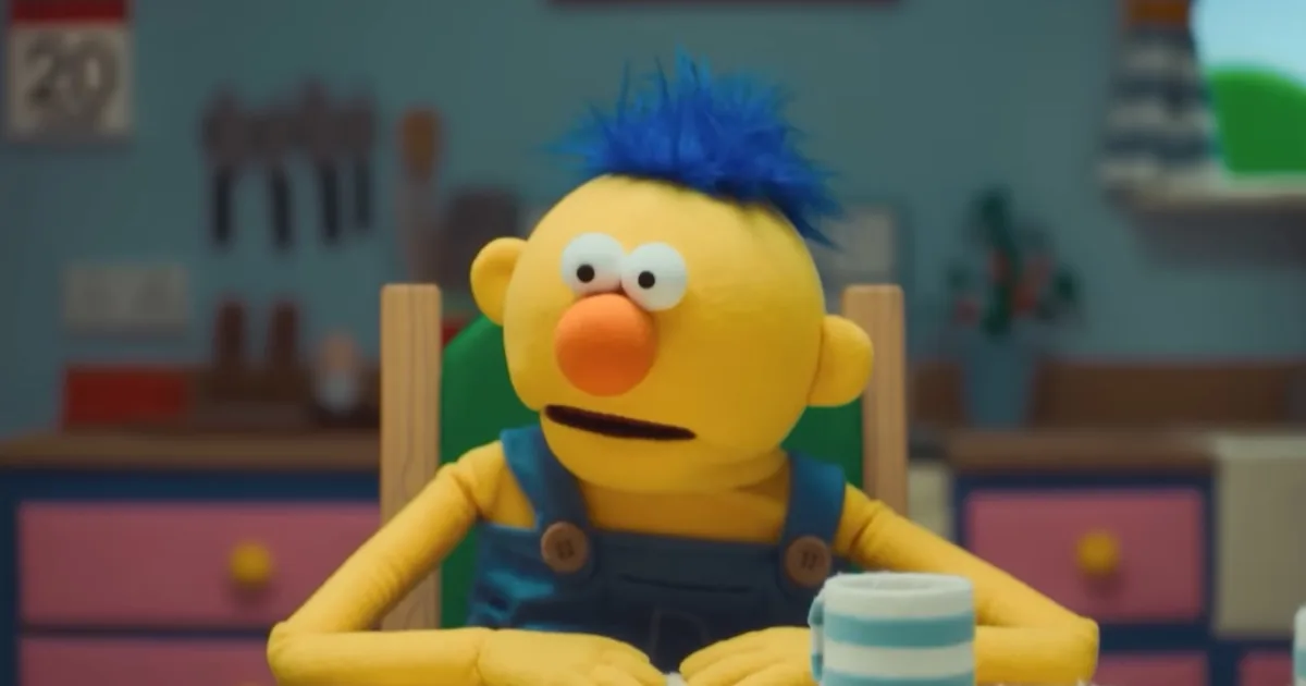 A yellow puppet with blue hair sits at a kitchen table.