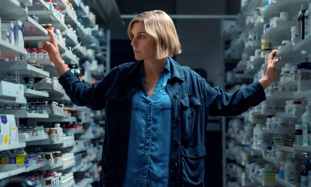 Pluribus1000x600 A woman with a blonde bob stands between two large shelving units filled with medications.