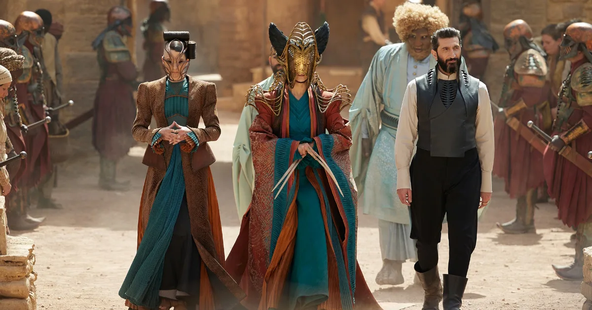 Women in elaborate gowns walk in a desert environment next to a man in a black vest.