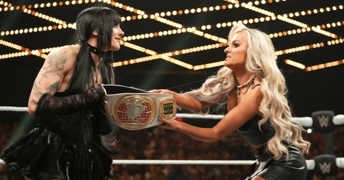 Two women hold a wrestling belt between them. It looks like the woman on the right is trying to take it from the woman on the left.