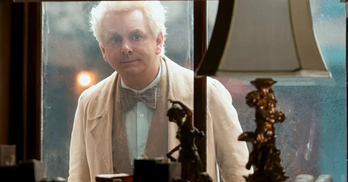 Aziraphale, a man with white hair and a bow tie, looks sadly into the window of a bookshop.