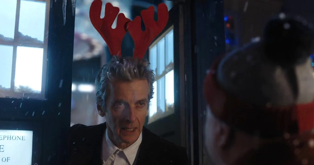 Peter Capaldi stands in the doorway of the TARDIS, wearing a pair of felt reindeer antlers.