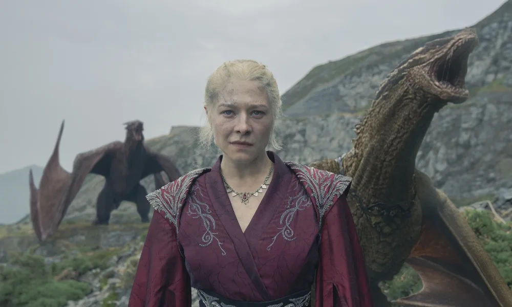 Syrax1000x600 A woman stands in front of two dragons.