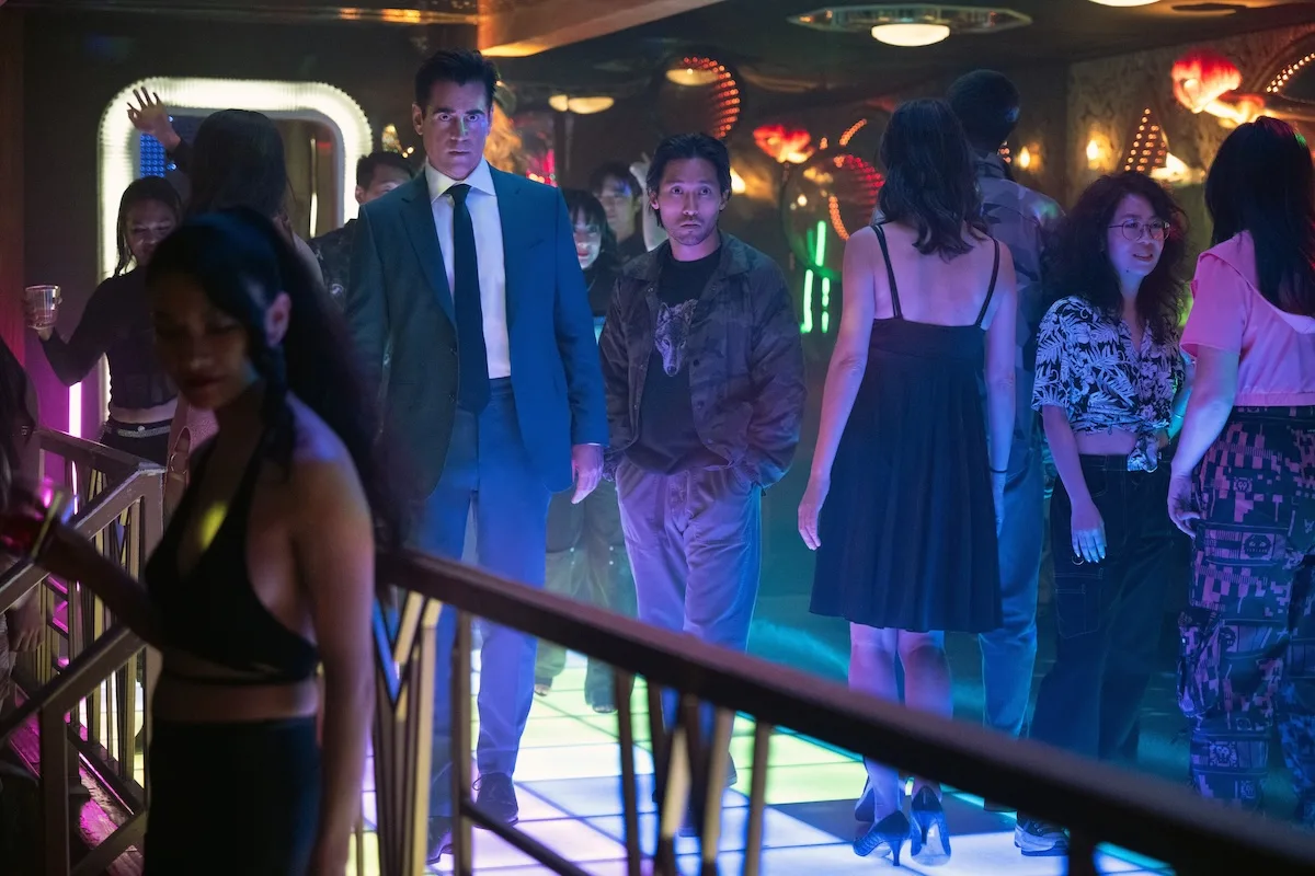 People mingle in a club while two men look intently at something offscreen.