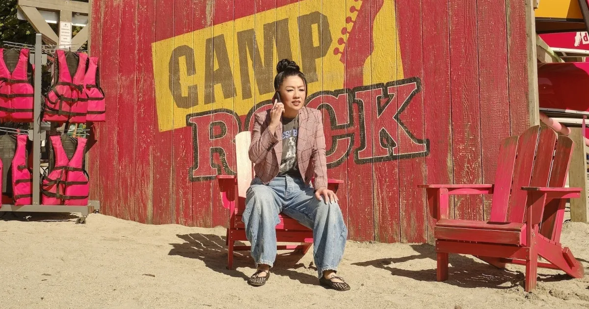 CampRock3_1200x600 A woman is on the phone, sitting in a red Adirondack chair in front of a sign that says "Camp Rock."