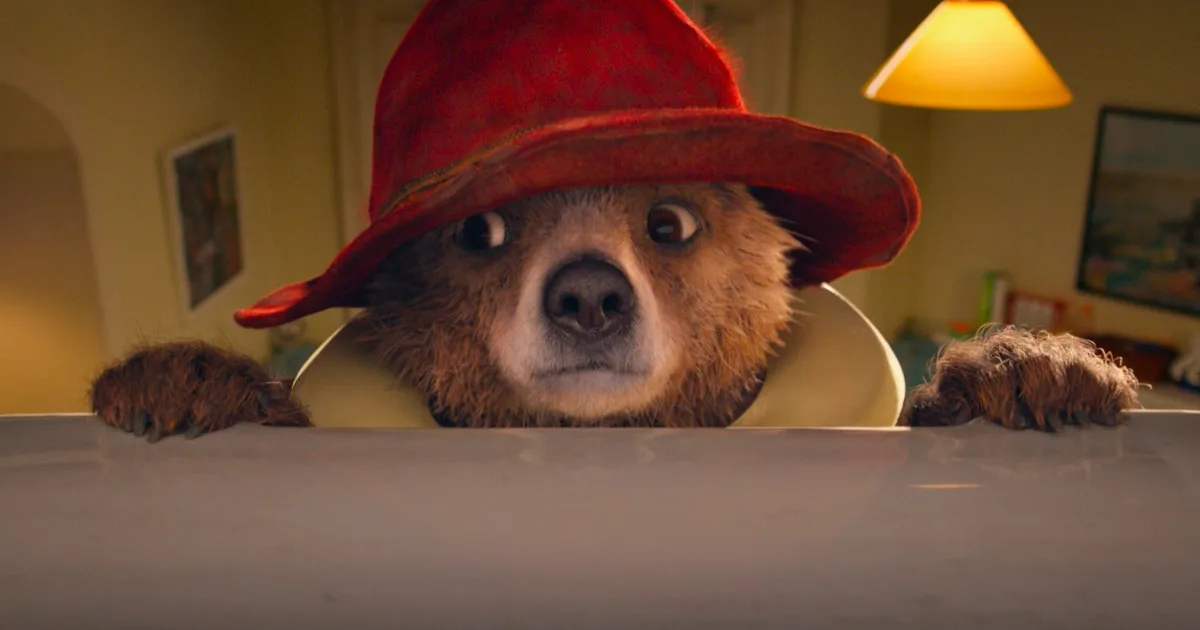 Paddington, a little CGI bear wearing a red hat, holds onto a ledge with his paws.