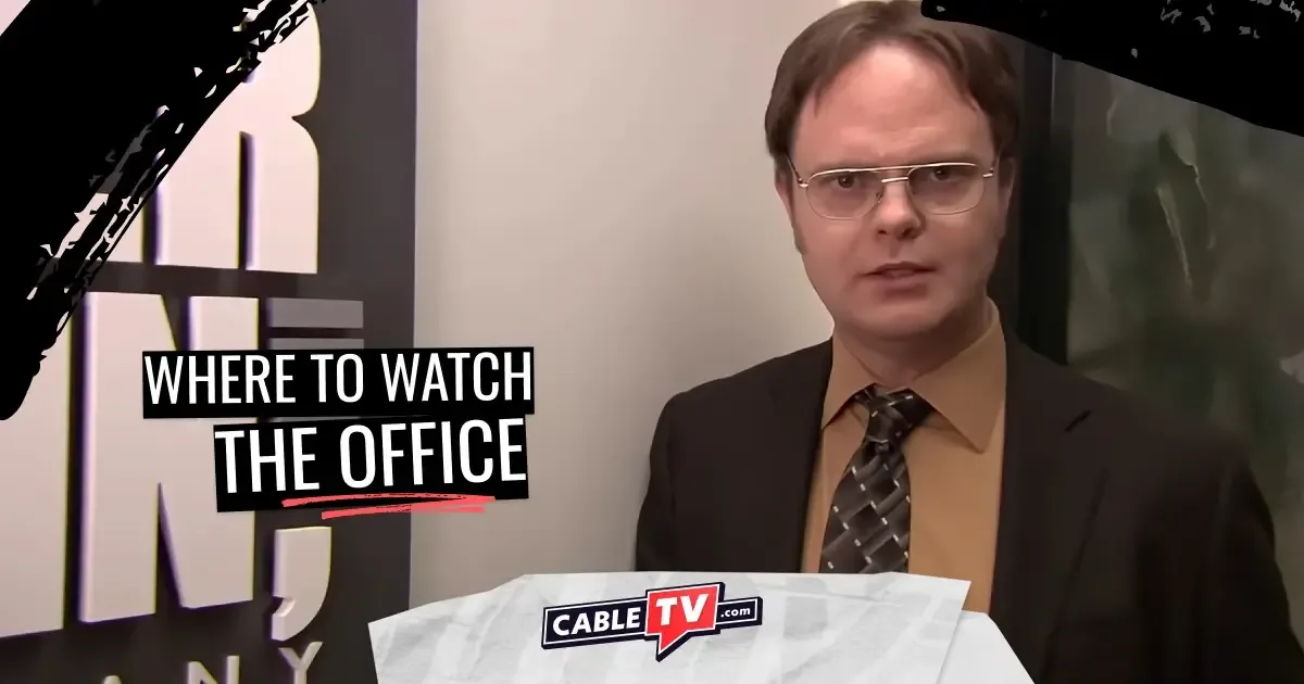 In an office, a man in a brown suit and glasses talks to the camera.