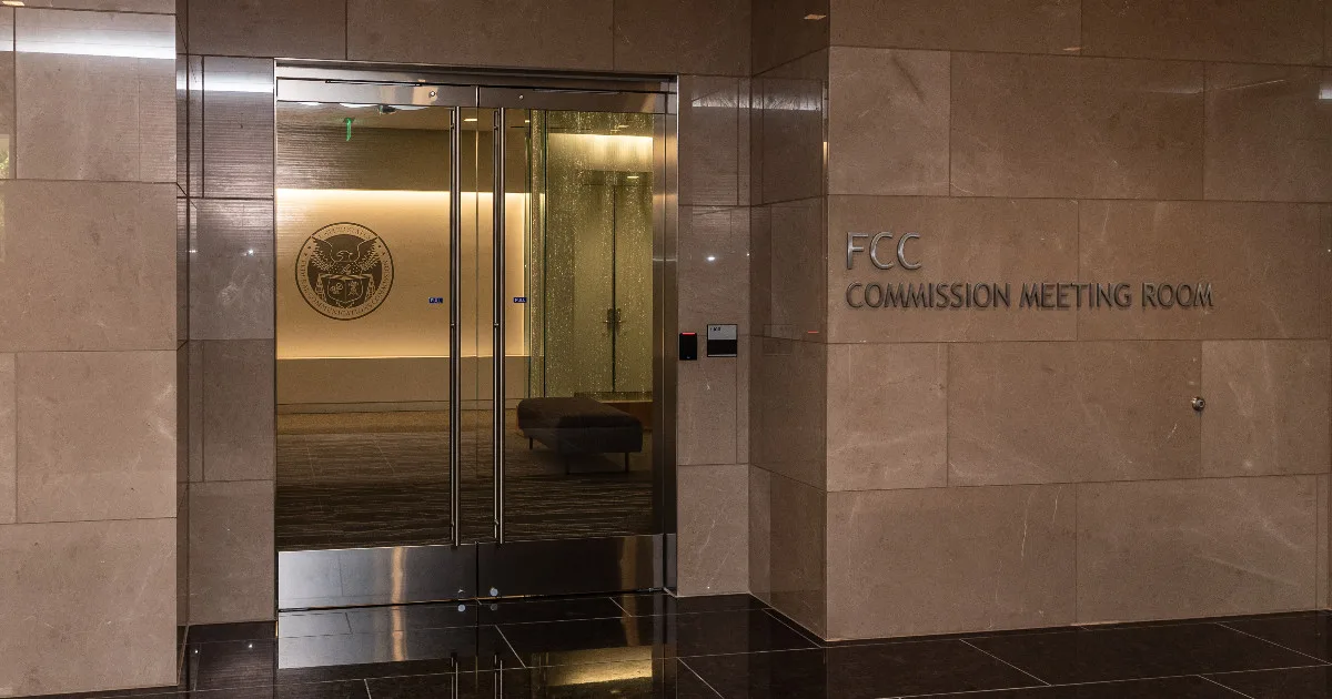 A photograph of the outside of the FCC Commission Meeting Room. There are a pair of glass double doors leading to a room with an FCC crest.