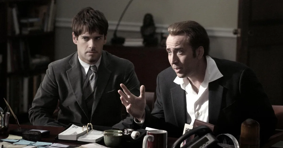 Two men sit at a desk. One of them is Nicolas Cage, explaining something to someone offscreen.