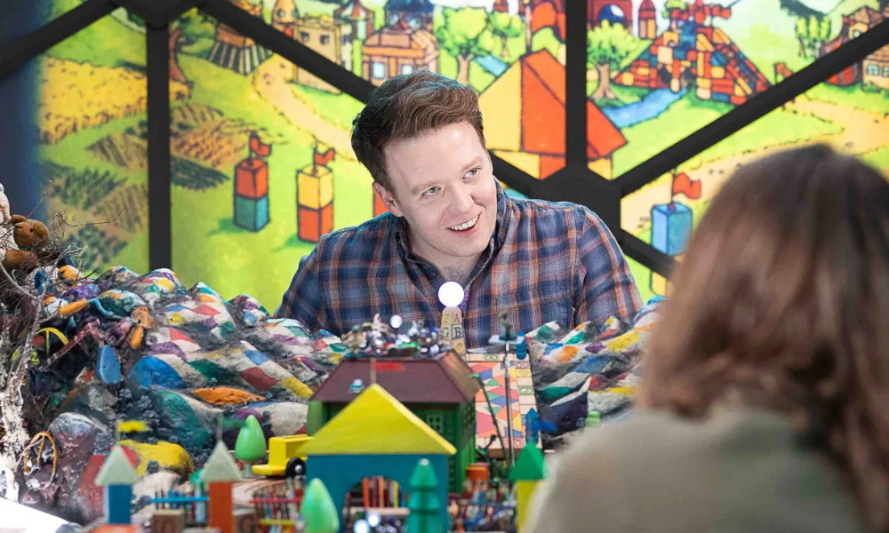 Brennan Lee Mulligan sits in the DM seat in the dome, surrounded by building-block castle imagery. He is smiling at a player.