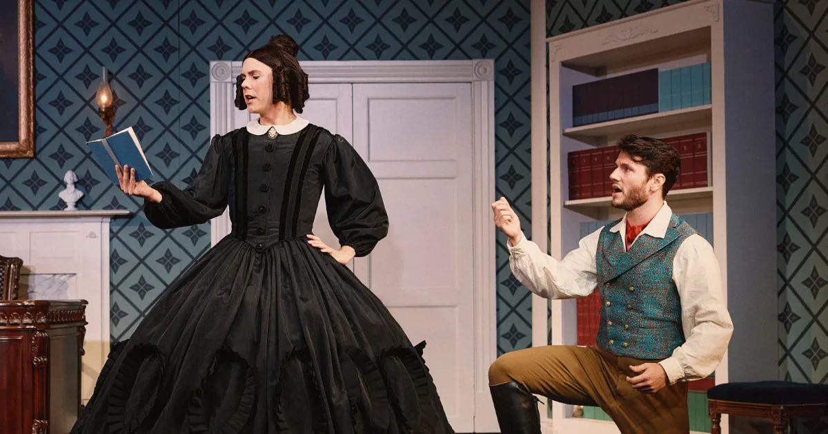 Cole Escola as Mary Todd Lincoln, reading a book in front of a man in a waistcoat.