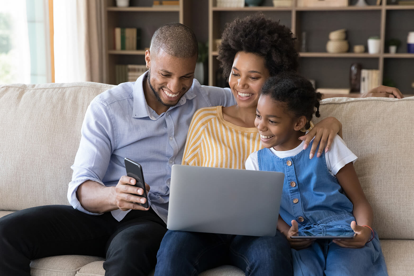 Happy African American parents and daughter resting on couch together watching online content on a laptop and mobile phone.